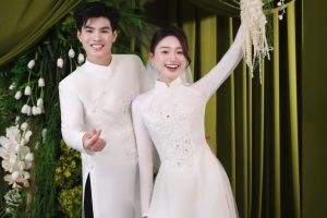 Cặp đôi mặc áo dài cưới trắng tinh khôi tạo dáng duyên dáng trước phông nền xanh sang trọng.