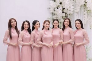 Một nhóm phụ nữ mặc áo dài màu hồng pastel chụp ảnh tại studio với hoa trắng.