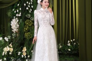 Cô dâu mặc váy cưới ren trắng sang trọng tạo dáng trước phông nền hoa và rèm xanh.